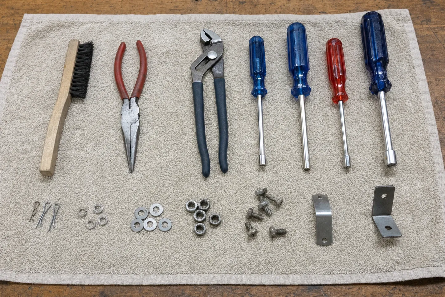Wire brush, two pairs of pliers and four nut drivers laid out on a towel with cotter pins, washers, nuts and brackets