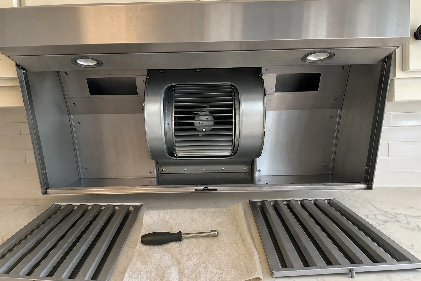 Stainless canopy hood with its filter panel removed, the twin squirrel-cage blower exposed inside, and two baffle filters lying on the counter below